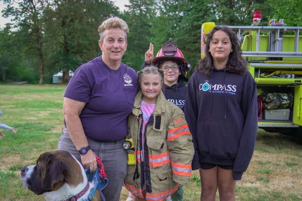 Three campers, one dressed in firefighter's gear, standing with a paramedic at a safety demonstration